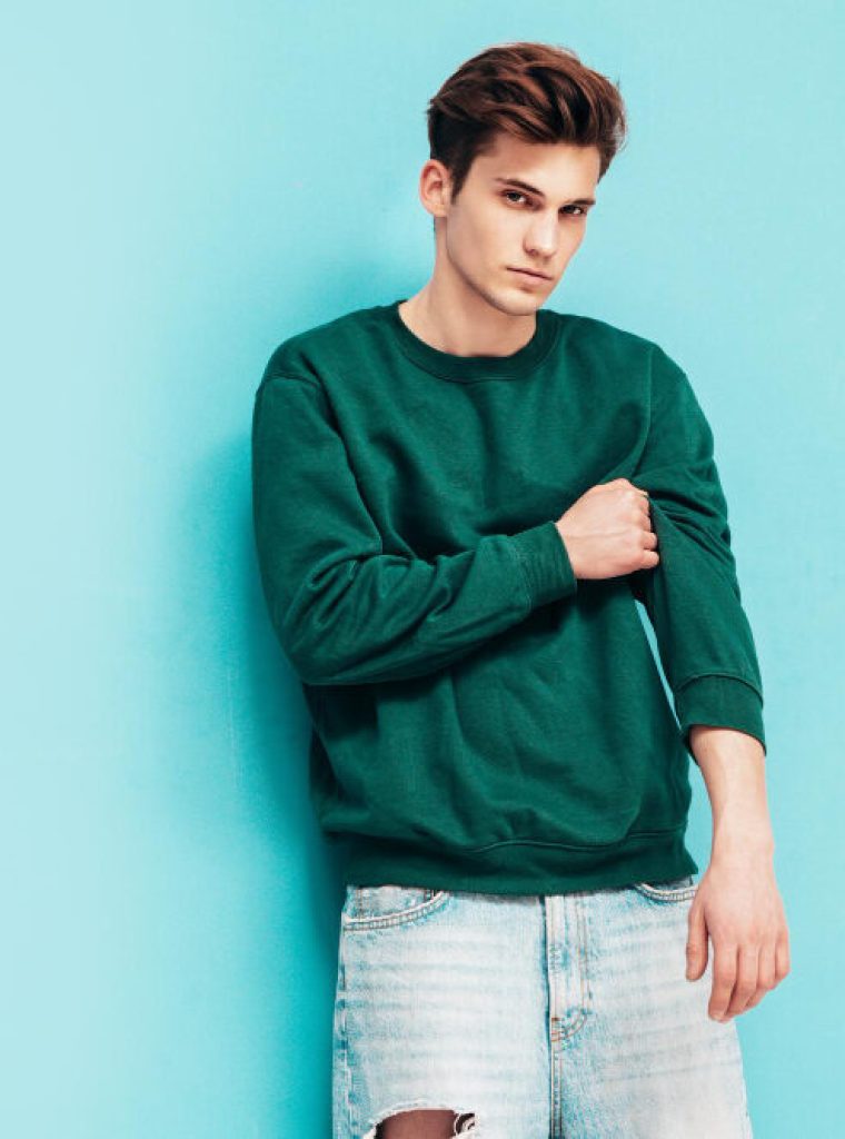 portrait-handsome-confident-stylish-hipster-lambersexual-modelman-dressed-green-sweater-jeans-fashion-male-posing-studio-near-blue-wall